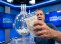 Homem de meia-idade segura dois bilhetes da Loteria dos Sonhos diante de globo com bolas brancas em estúdio azul