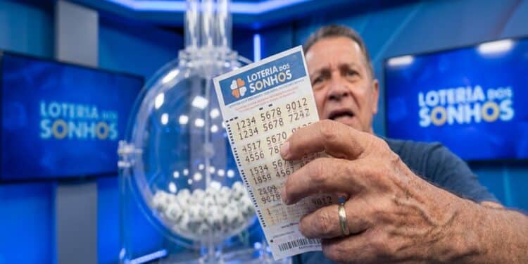 Homem de meia-idade segura dois bilhetes da Loteria dos Sonhos diante de globo com bolas brancas em estúdio azul