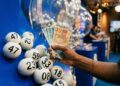 Mão segurando notas de 50 e 100 reais ao lado de bolas brancas numeradas em globo de sorteio azul