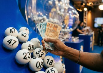 Mão segurando notas de 50 e 100 reais ao lado de bolas brancas numeradas em globo de sorteio azul