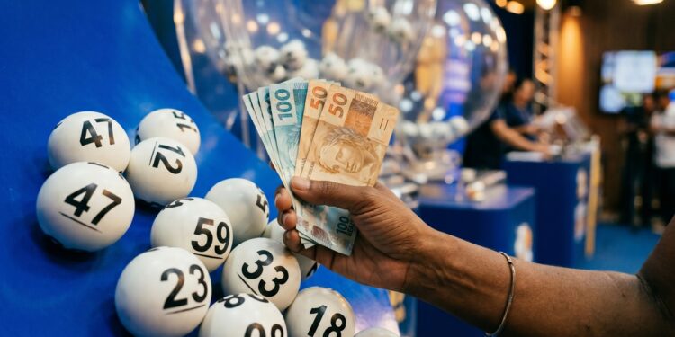 Mão segurando notas de 50 e 100 reais ao lado de bolas brancas numeradas em globo de sorteio azul