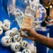 Mão segurando notas de 50 e 100 reais ao lado de bolas brancas numeradas em globo de sorteio azul