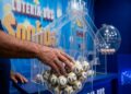 Mão masculina pegando bolas de sorteio douradas e brancas numeradas em globo de acrílico da Loteria dos Sonhos