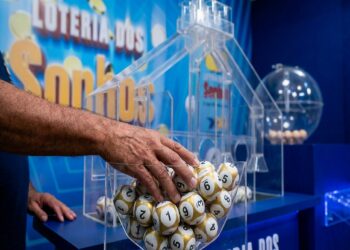 Mão masculina pegando bolas de sorteio douradas e brancas numeradas em globo de acrílico da Loteria dos Sonhos