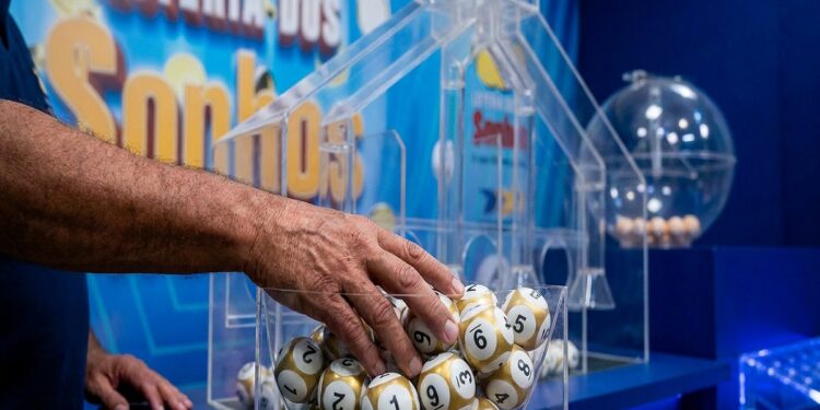 Mão masculina pegando bolas de sorteio douradas e brancas numeradas em globo de acrílico da Loteria dos Sonhos