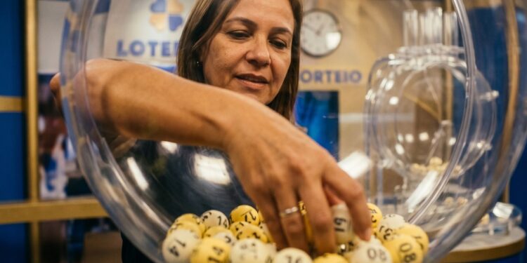 Mulher retira bola numerada de globo transparente em sorteio da Loteria do Ceará em Fortaleza, com painel azul ao fundo