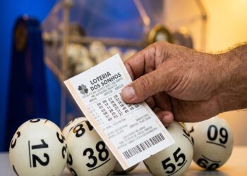 Mão segurando bilhete da Loteria dos Sonhos com bolas numeradas 12, 38, 09, 55 e 60 ao fundo