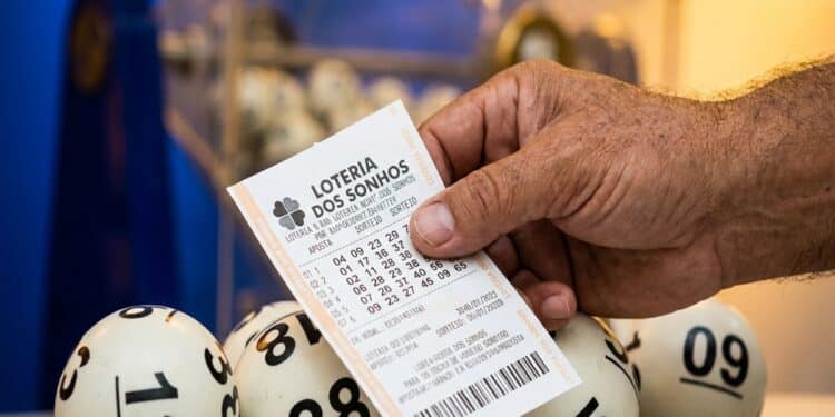 Mão segurando bilhete da Loteria dos Sonhos com bolas numeradas 12, 38, 09, 55 e 60 ao fundo