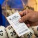 Mão segurando bilhete da Loteria dos Sonhos com bolas numeradas 12, 38, 09, 55 e 60 ao fundo
