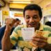 Homem de meia-idade comemora com punho cerrado segurando bilhete da Loteria dos Sonhos e notas de real em casa lotérica