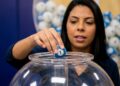 Mulher segura bola azul com número 1 sobre globo de acrílico com bolas coloridas ao fundo em sorteio