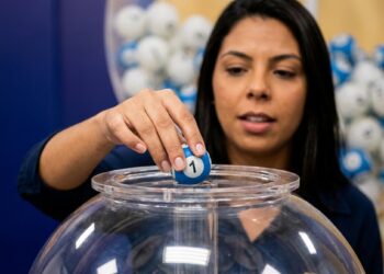 Mulher segura bola azul com número 1 sobre globo de acrílico com bolas coloridas ao fundo em sorteio