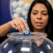 Mulher segura bola azul com número 1 sobre globo de acrílico com bolas coloridas ao fundo em sorteio