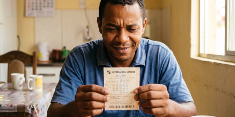 Homem negro de camisa polo azul sorrindo ao conferir bilhete da Loteria dos Sonhos com data 21/05 em cozinha residencial