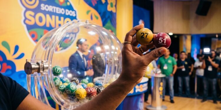Mão segura bolas amarelas com número 27 e vinho com número 51 durante sorteio da Loteria dos Sonhos no Ceará