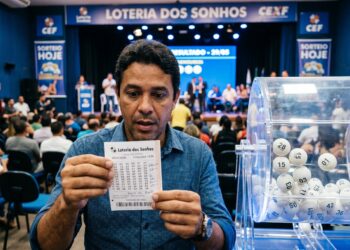 Homem de camisa azul segurando bilhete da Loteria dos Sonhos, com globo de sorteio e bolas numeradas à direita