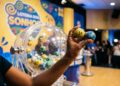 Mão segurando bolas amarela (14) e azul (36) em sorteio da Loteria dos Sonhos, com globo de bolas ao fundo