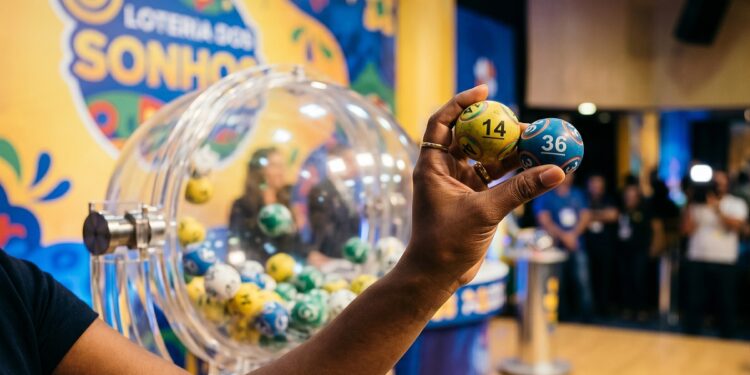 Mão segurando bolas amarela (14) e azul (36) em sorteio da Loteria dos Sonhos, com globo de bolas ao fundo
