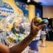 Mão segurando bolas amarela (14) e azul (36) em sorteio da Loteria dos Sonhos, com globo de bolas ao fundo