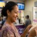 Mulher sorrindo segura volante da Lotofácil em agência Loterias Caixa, com painéis de resultados ao fundo