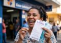 Mulher sorridente segura volante da Lotofácil e notas de R$ 100 diante da agência da Caixa Econômica Federal