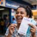 Mulher sorridente segura volante da Lotofácil e notas de R$ 100 diante da agência da Caixa Econômica Federal