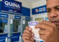 Homem de meia-idade segura e observa bilhete da Quina em frente ao guichê de lotérica da Caixa
