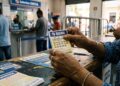 Mãos segurando bilhete da Quina, concurso 7034, com números apostados em balcão de Loterias Caixa