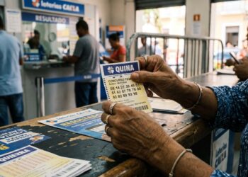 Mãos segurando bilhete da Quina, concurso 7034, com números apostados em balcão de Loterias Caixa
