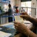 Mãos segurando bilhete da Quina, concurso 7034, com números apostados em balcão de Loterias Caixa
