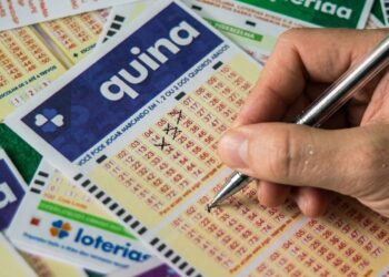 Mão segurando caneta marca números em volante da Quina, com bilhetes das Loterias Caixa ao fundo