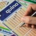 Mão segurando caneta marca números em volante da Quina, com bilhetes das Loterias Caixa ao fundo