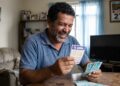 Homem sorridente segura volante da Quina e notas de R$ 100 em sala de estar com calendário marcando 27 de maio