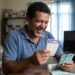 Homem sorridente segura volante da Quina e notas de R$ 100 em sala de estar com calendário marcando 27 de maio