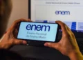 Mãos segurando smartphone com logo do ENEM na tela, em frente a monitor de computador exibindo a Página do Participante.