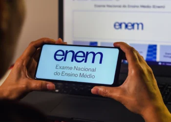 Mãos segurando smartphone com logo do ENEM na tela, em frente a monitor de computador exibindo a Página do Participante.