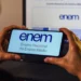 Mãos segurando smartphone com logo do ENEM na tela, em frente a monitor de computador exibindo a Página do Participante.