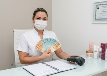 Profissional de estética com máscara branca segura notas de R$100 e R$50 em leque ao lado de maquininha de cartão.
