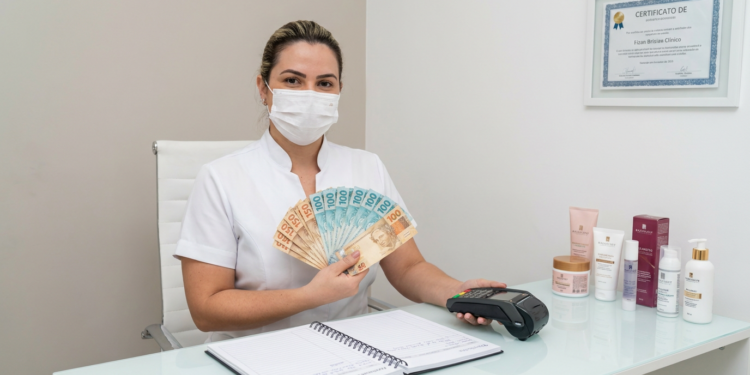Profissional de estética com máscara branca segura notas de R$100 e R$50 em leque ao lado de maquininha de cartão.