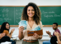 Professora de cabelos cacheados sorri segurando notas de R$ 100 e carteira marrom enquanto alunos aplaudem em sala de aula.
