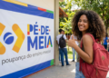 Jovem de cabelos cacheados com mochila vermelha faz sinal de positivo ao lado do banner Pé-de-Meia em escola pública.