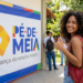 Jovem de cabelos cacheados com mochila vermelha faz sinal de positivo ao lado do banner Pé-de-Meia em escola pública.