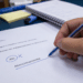 Mão segurando caneta azul sublinhando "Atenciosamente" em carta comercial brasileira sobre mesa com caderno e caneca.