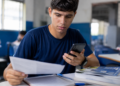Jovem de camiseta azul-marinho sentado em sala de aula segura folha de papel na mão direita e smartphone na esquerda.
