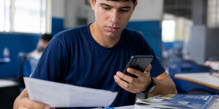 Jovem de camiseta azul-marinho sentado em sala de aula segura folha de papel na mão direita e smartphone na esquerda.