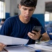 Jovem de camiseta azul-marinho sentado em sala de aula segura folha de papel na mão direita e smartphone na esquerda.