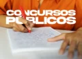 Pessoa de camisa laranja escreve com caneta vermelha em caderno sobre mesa, com texto "Concursos Públicos".