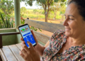 Mulher sorri ao acessar o aplicativo do Bolsa Família no celular, sentada em banco de madeira ao ar livre.