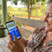 Mulher sorri ao acessar o aplicativo do Bolsa Família no celular, sentada em banco de madeira ao ar livre.