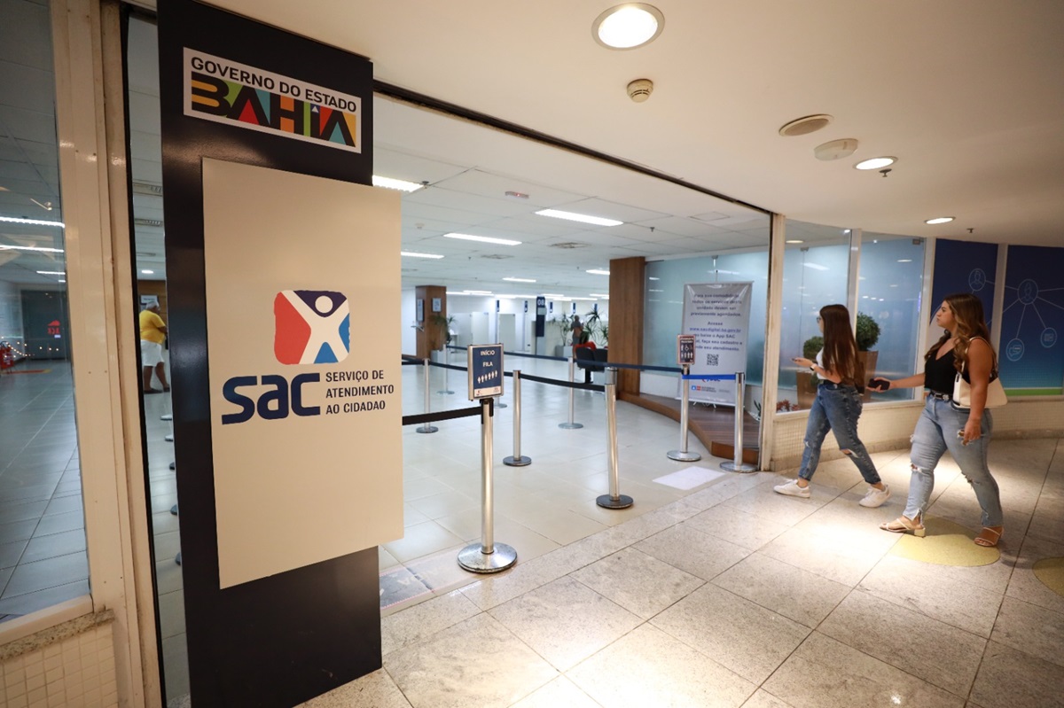 Entrada de uma unidade do SAC em corredor de shopping com totem do Governo do Estado da Bahia e duas mulheres caminhando ao lado